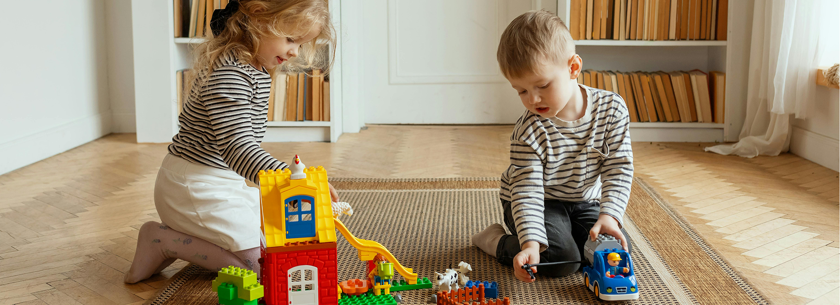 Dziewczynka i chłopiec siedzą na dywanie i bawią się dużymi klockami Lego Duplo. Budują domek ze zjeżdżalnią, a obok stoją figurki zwierząt. Chłopiec trzyma w ręce samochód. W tle widać białe regały z książkami.