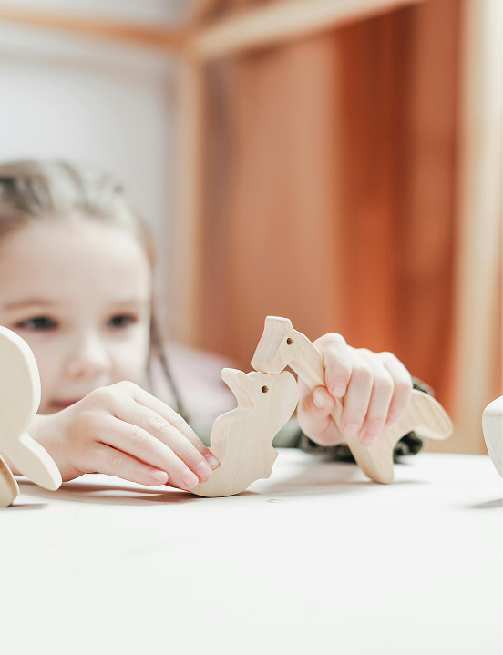 Mała dziewczynka bawi się przy białym stole dwoma drewnianymi figurkami dinozaurów. Jej dłonie trzymają dinozaury blisko siebie, imitując interakcję. Inne drewniane zabawki leżą na stole.