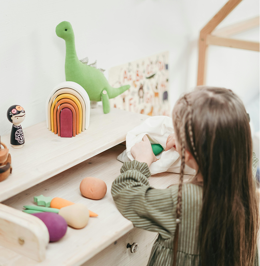 Dziewczynka z zaplecionymi warkoczykami na głowie kładzie pluszowego dinozaura obok drewnianych warzyw na komodzie.