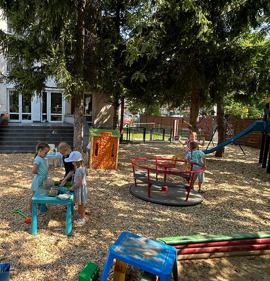 Przestronny plac zabaw znajdujący się wśród drzew, które dają dużo cienia. Dzieci bawią się przy stoliku oraz na karuzeli. W tle widoczny budynek przedszkola z napisem "Przedszkole Czyżyk".