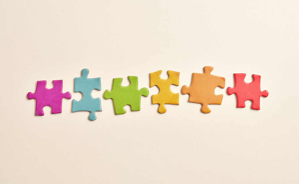 Sześć pojedynczych, różnokolorowych elementów puzzli ułożonych obok siebie na jasnobeżowym tle. Elementy są w kolorach: fioletowym, jasnoniebieskim, jasnozielonym, żółtym, pomarańczowym oraz czerwonym.
