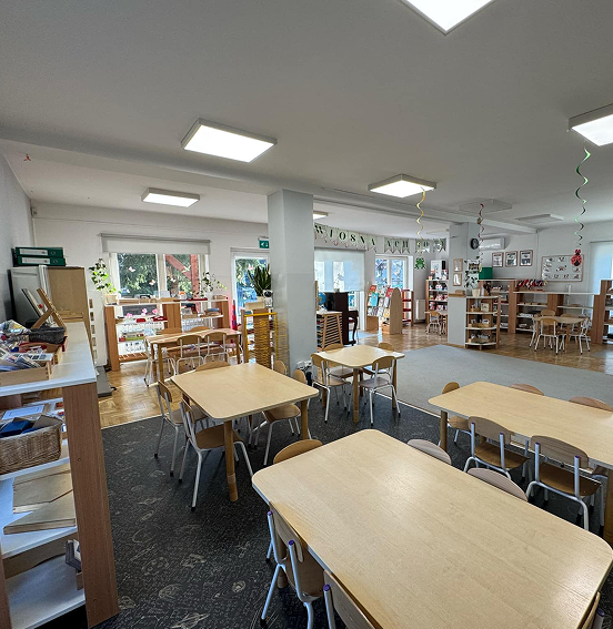 Kącik lekcyjny z małymi stolikami z krzesełkami, niskimi regałami z materiałami edukacyjnymi i zabawkami. Na ścianach widać wiosenne dekoracje z papieru.
