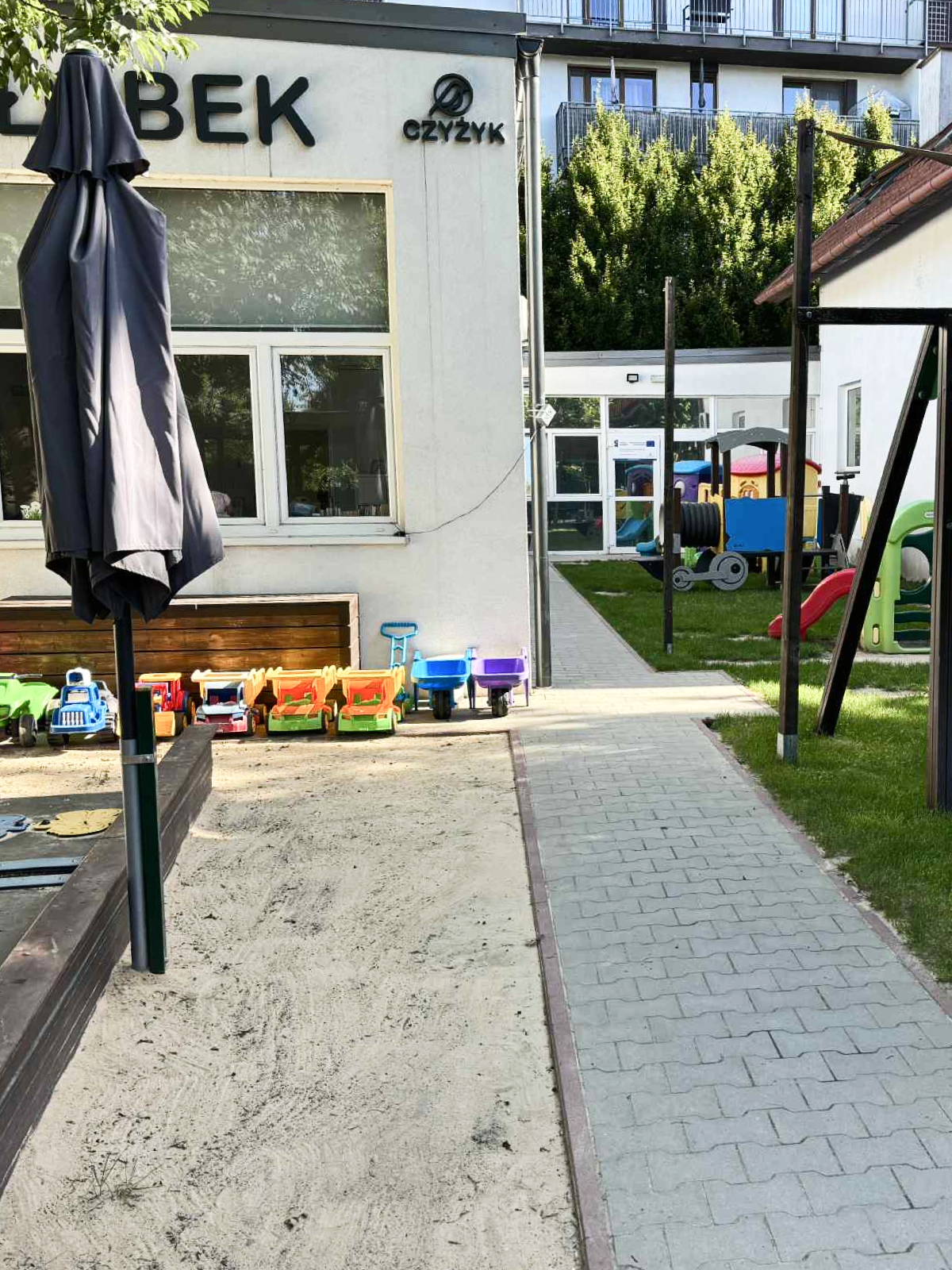 Plac zabaw z różnymi zabawkami oraz huśtawką, zjeżdżalnią i ciuchcią. Na piasku znajduje się złożony parasol, a stoliki znajdują się pod daszkiem.